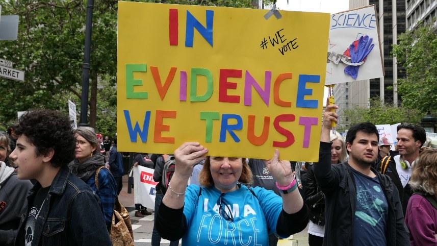Woman holding an "In Evidence We Trust" poster.